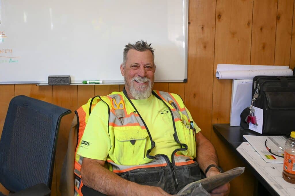 A smiling electrical contractor in Boise, Idaho, in a neon yellow safety shirt and vest, sits in a wood-paneled office with work papers.